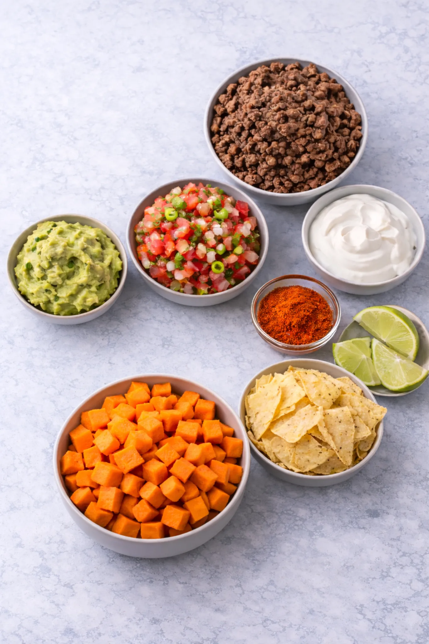 Sweet potato taco bowl ingredients arranged in bowls with roasted sweet potatoes, ground beef, guacamole, pico de gallo, sour cream, taco seasoning, tortilla chips, and lime wedges