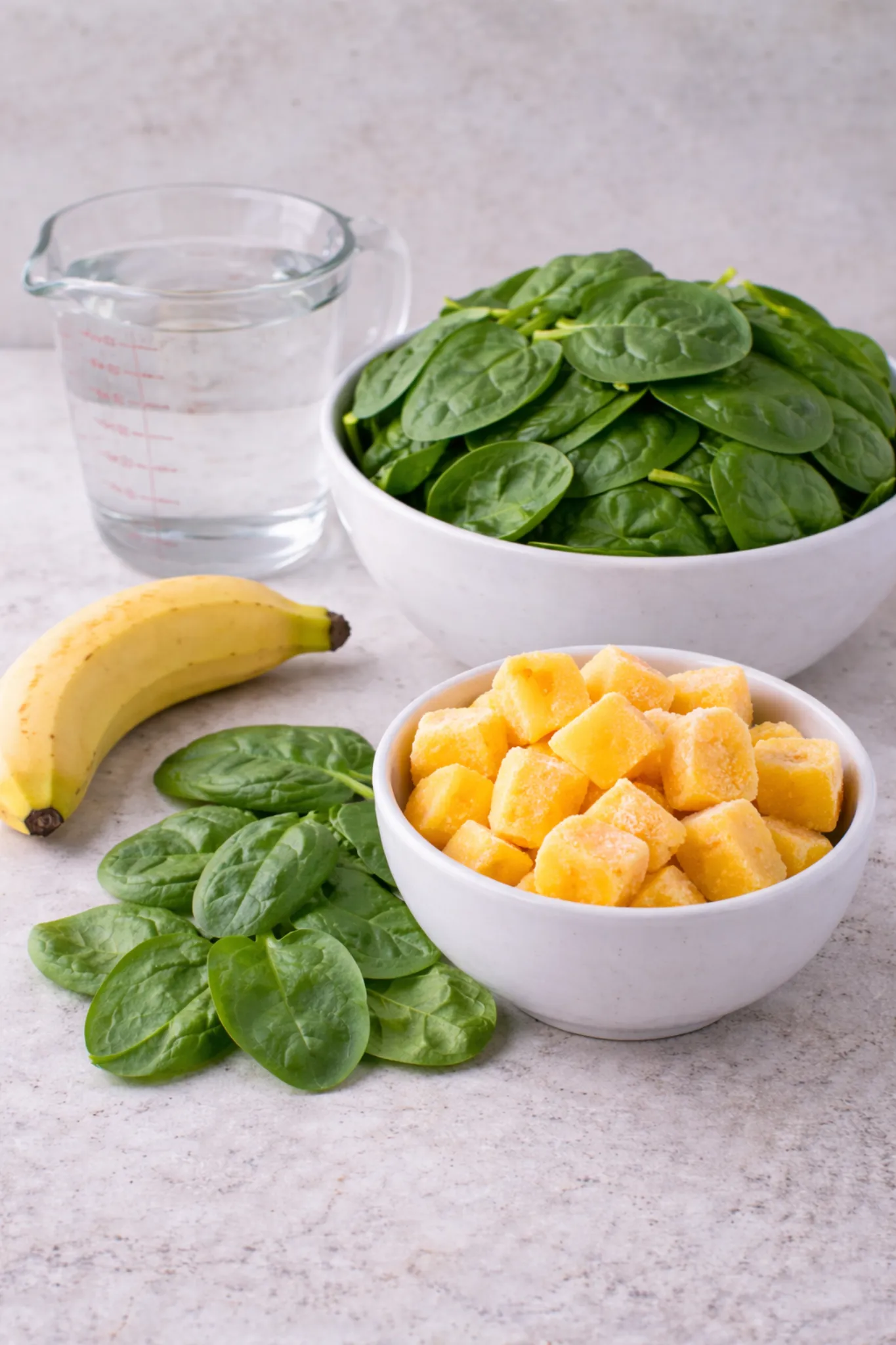 Ingredients for a green smoothie: fresh spinach, banana, frozen mango chunks and water on a light background